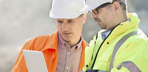 Zwei Männer mit einem Bauhelm auf dem Kopf gucken auf einem Laptop